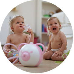Two happy baby girls sitting on the floor with the Nosiboo Pro Electric Nasal Aspirator