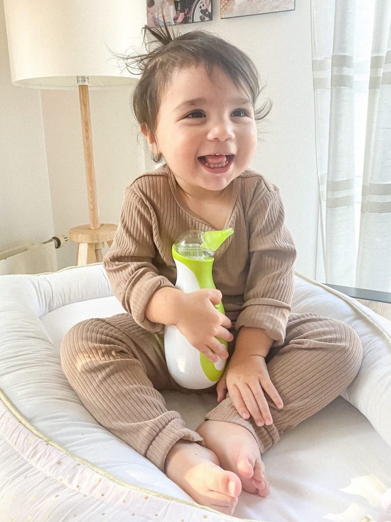 Smiling little girl in a baby nest holding a Nosiboo Go portable electric nasal aspirator